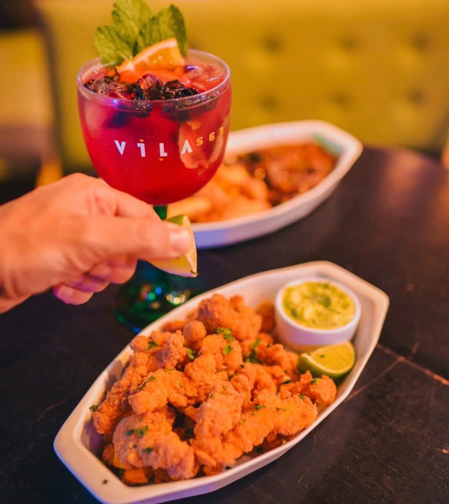 Porção de frango empanado com guacamole e limão, ao lado de um drink do Vila